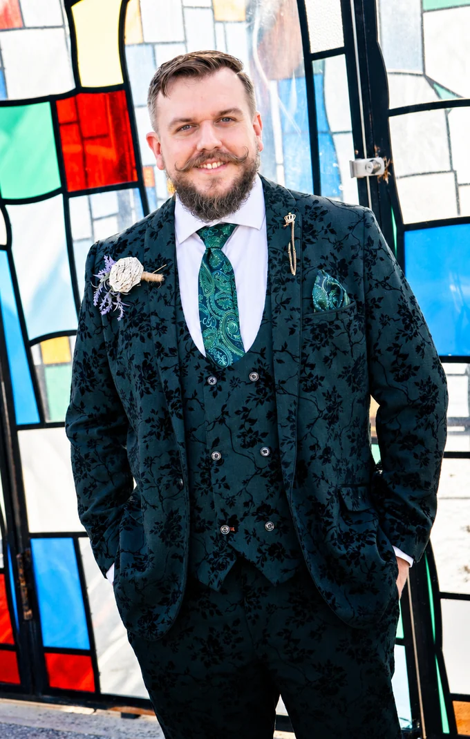 Michael Nault, standing in front of a stained-glass door in a dark green floral three-piece suit with a teal paisley tie and pocket square.